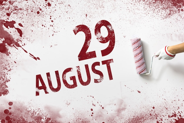 August 29th. day 29 of month, calendar date. the hand holds a\
roller with red paint and writes a calendar date on a white\
background. summer month, day of the year concept.