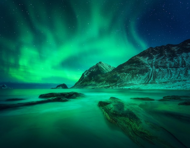 Aurora borealis boven besneeuwde berg en zandstrand met stenen Noorderlicht op de Lofoten-eilanden Noorwegen Sterrenhemel met poollicht Nacht winterlandschap met aurora zee met wazig water