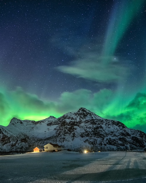 Aurora boreale con stellato sulla montagna innevata e villaggio nordico di notte a flakstad, isole lofoten, norvegia