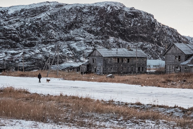 Authentic Russian northern village, old dilapidated wooden houses, harsh Arctic nature. A man walks with a dog.