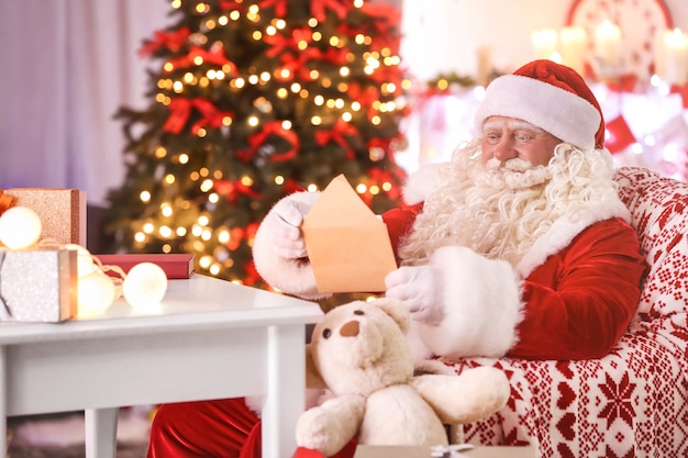 Authentic Santa Claus reading letter at table in room decorated for Christmas