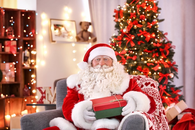 Authentic Santa Claus with gift box sitting in armchair at room decorated for Christmas