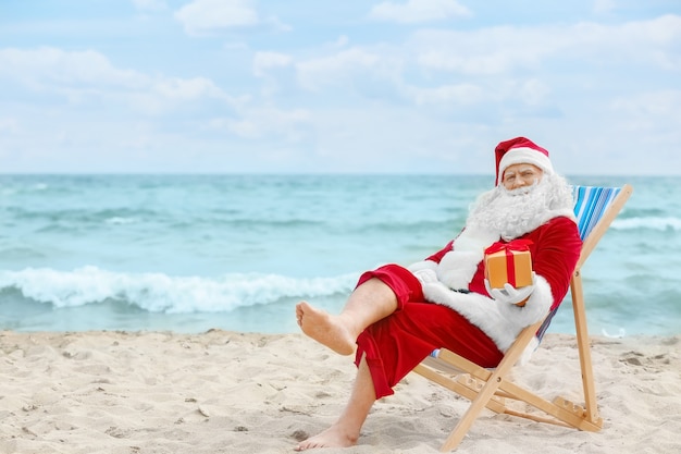 Authentieke kerstman met geschenkdoos in ligstoel op strand