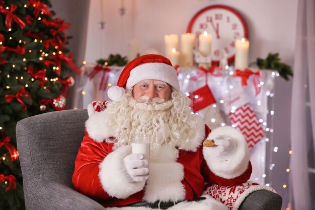 Authentieke Kerstman met koekje en glas melk zittend in een stoel op kamer ingericht voor Kerstmis