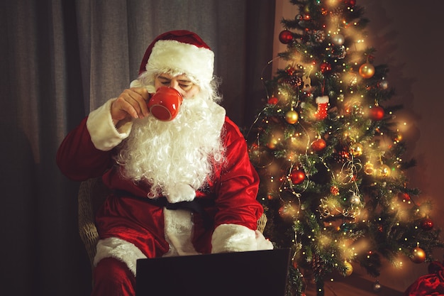 Authentieke kerstman werkt achter een laptop en drinkt warme chocolademelk. Huis interieur. In afwachting van Kerst en Oud en Nieuw.