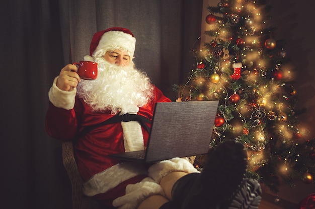 Authentieke Kerstman werkt op een laptop. Huis interieur. In afwachting van Kerst en Oud en Nieuw.