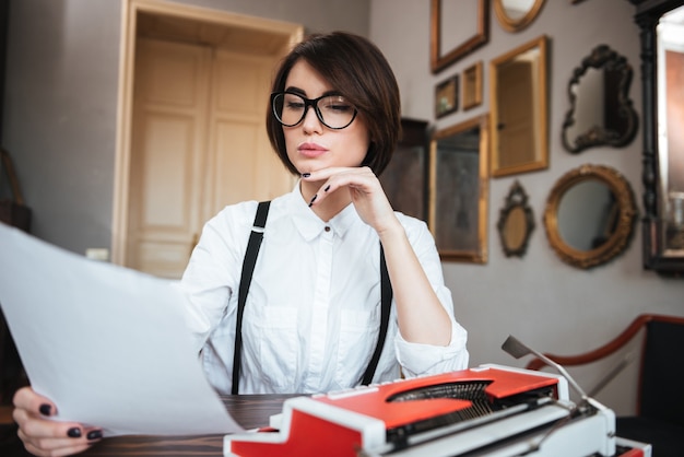 Authoress in glasses and white shirt sitting by the table with paper and typewriter