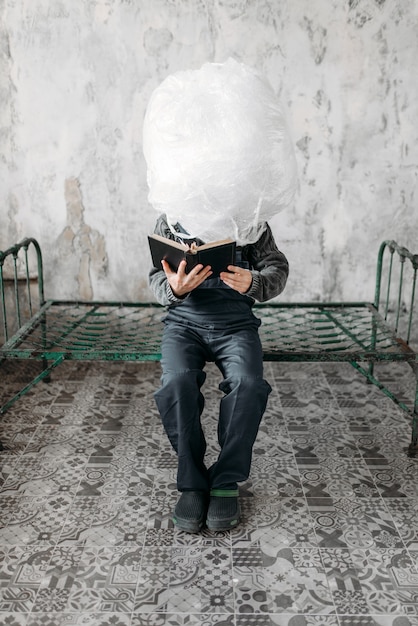 Autistic wraps his head in packaging film and sitting on the bed, grunge room interior.