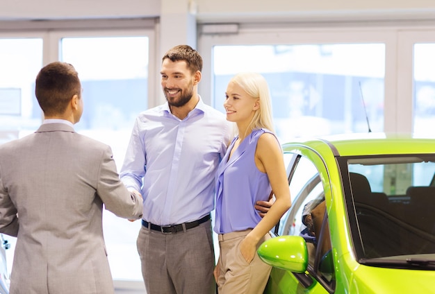 Фото Автомобильный бизнес, продажа автомобилей и концепция людей - счастливая пара с дилером пожимает руку в автосалоне или салоне