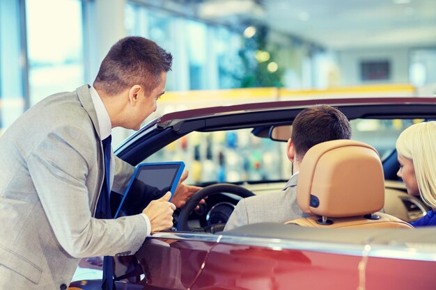 Auto business, car sale, consumerism and people concept - happy couple sitting in cabriolet with car dealer in auto show or salon