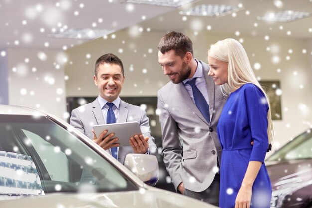 Photo auto business, car sale, technology and people concept - happy couple with car dealer in auto show or salon over snow effect