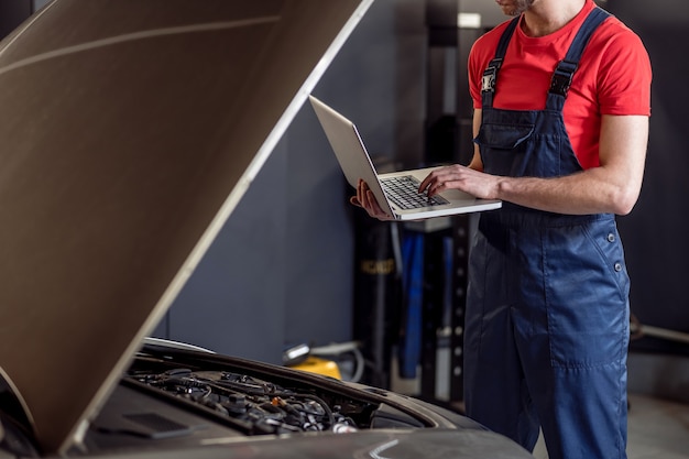 Auto, diagnostiek. Handen van automonteur in blauwe overall permanent met laptop in de buurt van open kap van auto in werkplaats