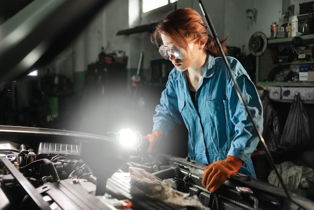 自動錠前屋の女の子がランプの光を照らしている車のエンジンを検査しますガレージまたは自動車修理店とオーバーオールと眼鏡で働いている女性機械修理の概念