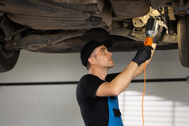 Auto mechanic in blue coveralls and black cap working underneath car lifting machine at the garage