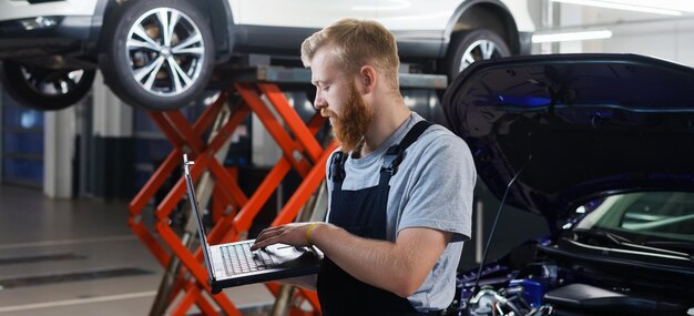 自動車整備士は、サービス ステーションを背景に専門的な診断用ラップトップを持って立っています。車両エラーの診断、パノラマ写真。