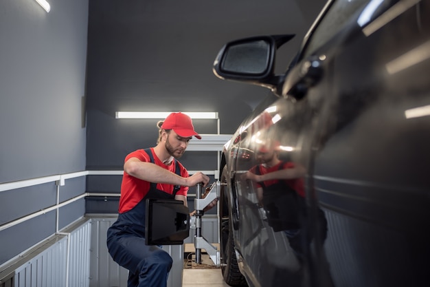 Auto repair shop. Young adult serious worker in overalls standing near wheel of car with special device