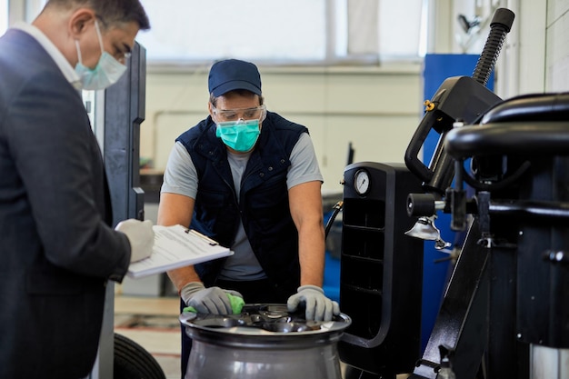 Riparatore automobilistico con maschera protettiva che esamina il pneumatico rotto dell'auto mentre il suo manager lo sta supervisionando in un'officina