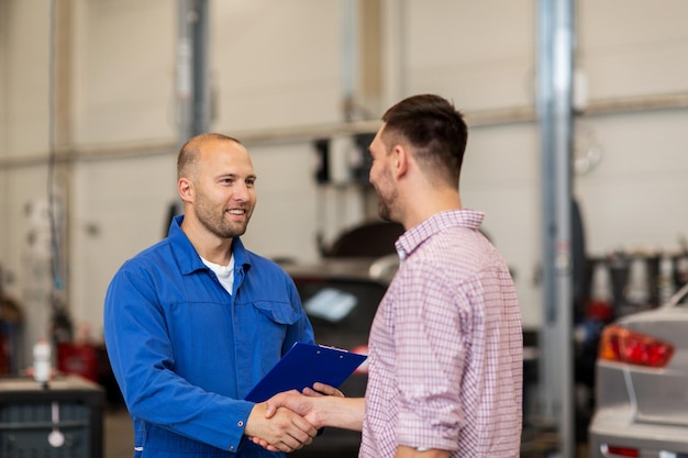 auto service, repair, maintenance, gesture and people concept - mechanic with clipboard and man or owner shaking hands at car shop