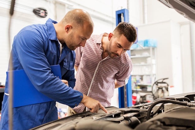 auto service, repair, maintenance and people concept - mechanic with clipboard and man or owner looking at broken car engine at shop