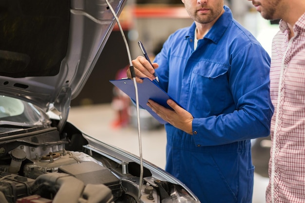 Photo auto service, repair, maintenance and people concept - mechanic with clipboard and man or owner looking at broken car at shop