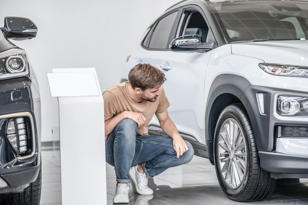 Auto wiel. Bezorgde gefocuste jongeman gehurkt in de buurt van het wiel van een nieuwe auto in een autodealer die overweegt