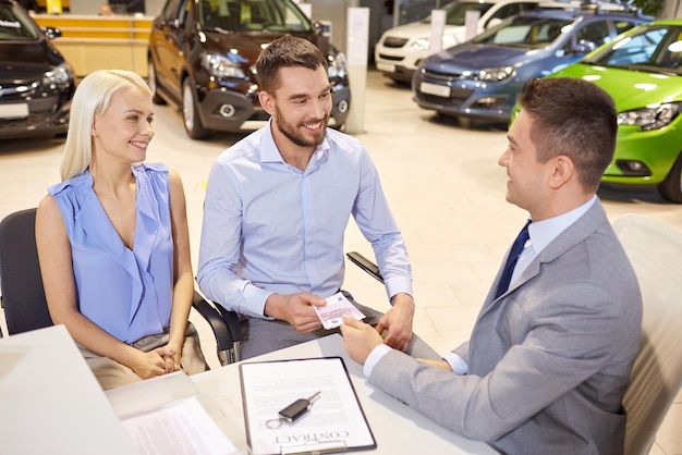 autobedrijf, verkoop en mensenconcept - gelukkig paar met dealer die auto koopt in autoshow of salon