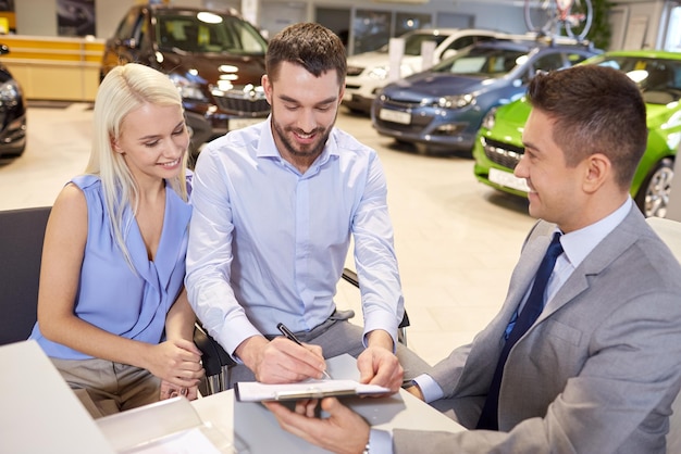 Foto autobedrijf, verkoop en mensenconcept - gelukkig stel met dealer die auto koopt en documenten ondertekent in autoshow of salon