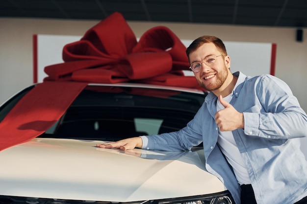 Automobile is covered in gift tape Successful man in glasses standing near brand new car indoors