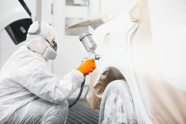 Маляр по ремонту автомобилей в маске и защитной спецодежде красит белый кузов автомобиля в покрасочной камере