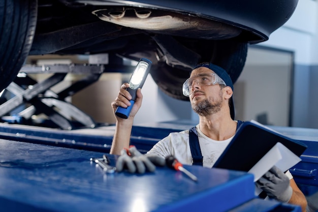 Automonteur die chassis van een voertuig onderzoekt met een zaklamp in een werkplaats