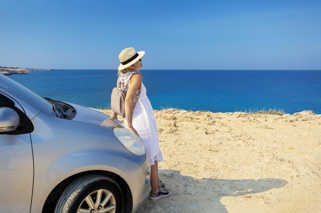 Autoreizen en vrijheidsconcept Gelukkige vrouwenreiziger op de strandweg met haar auto en overzeese strandachtergrond