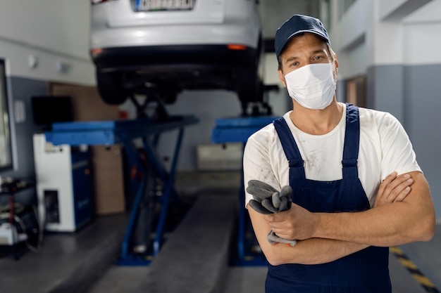 Autoreparateur staat met gekruiste armen terwijl hij een gezichtsmasker draagt in een werkplaats