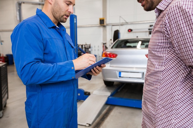 autoservice, reparatie, onderhoud en mensenconcept - monteur met klembord in gesprek met man of eigenaar in autowinkel