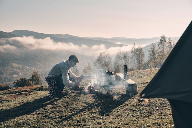 秋の冒険。山のテントの近くに座ってキャンプファイヤーを作る若い男