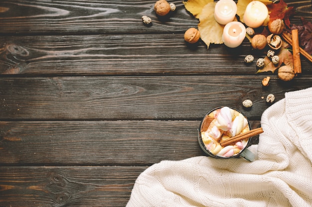 autumn background made of dried fall leaves, mug of cocoa with marshmallows, nuts, cinnamon, plaid, apples. top view on brown wood