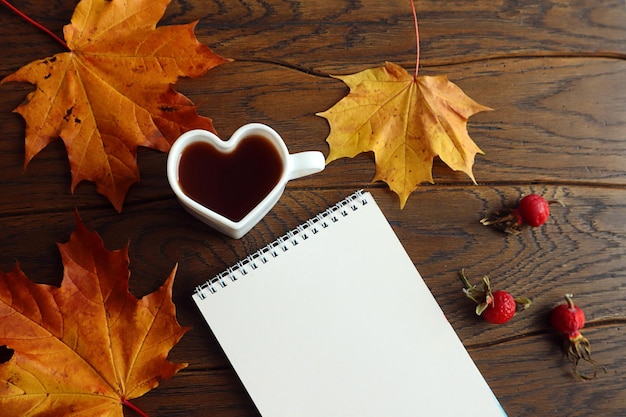 Autumn background notebook heartshaped cup of hot tea yellow maple leaves red rose hips on a wooden background top view space for text