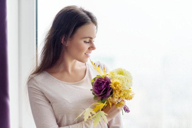 秋の背景、黄色の落ち葉と秋の花の花束が曇った雨の窓で見ている若い美しい女性、テキストのコピースペース