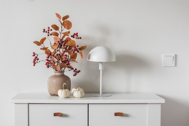 Autumn bouquet in a ceramic vase scandinavian style table lamp decorative pumpkins on a white chest of drawers