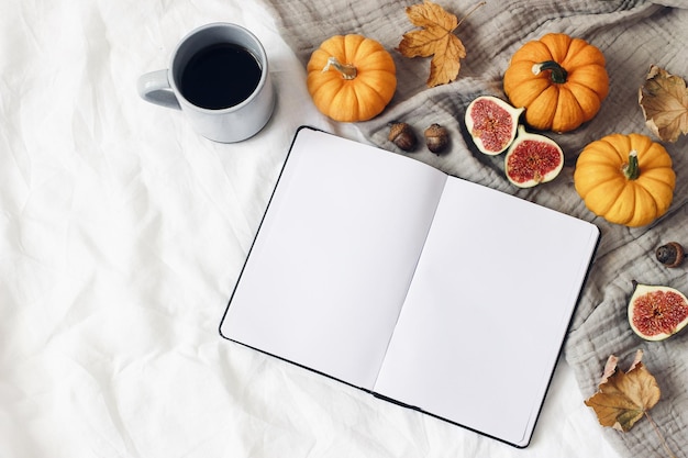 Autumn breakfast in bed Blank open notebook mockup Cup of coffee colorful leaves figs and little orange pumpkins White linen background Thanksgiving halloween concept Flat lay top view