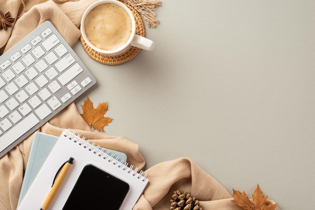 Autumn business concept Top view photo of notepads pen smartphone keyboard cup of coffee on rattan placemat anise yellow maple leaves pine cone and scarf on isolated grey background