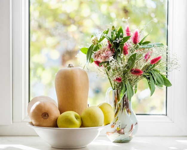 Autumn composition bouquet of flowers in a vase butternut squashes apples in a bowl against window pane