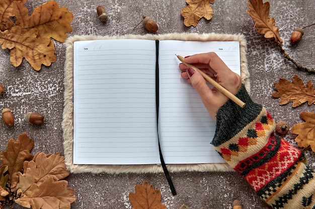 Autumn composition made of brown dried leaves and acorns on dark concrete background. Template mockup blank notebook and hand with a pen. Fall, halloween. Flat lay, copy space background.