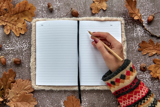 Foto composizione autunnale fatta di foglie secche marroni e ghiande su fondo di cemento scuro. taccuino in bianco del modello del modello e mano con una penna. autunno, halloween. lay piatto, copia dello sfondo dello spazio.