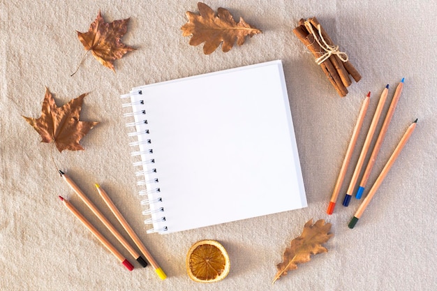 Фото Осенняя композиция с пустым альбомом для рисования, цветные карандаши, осенние сухие листья, сушеная апельсиновая корица на фоне льняной ткани, плоская планировка, копия пространства, осенняя концепция