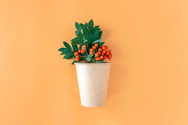Composizione autunnale con bicchiere di carta ecologico e mazzo di sorbo su sfondo arancione. natura morta autunnale minimalista, elegante e creativa. lay piatto, copia spazio.