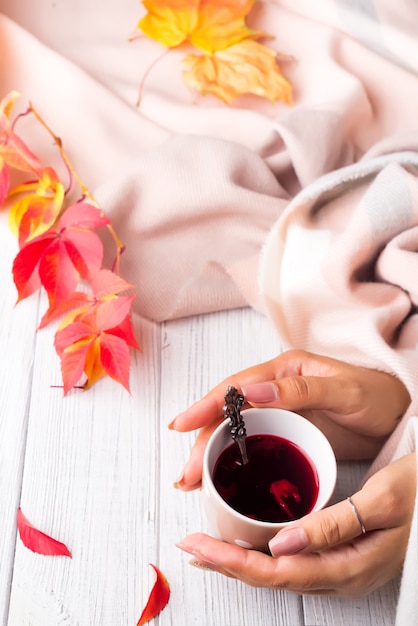 Autumn concept. Woman hands hold A cup of healthy tea in yellow red leaves with beige scarf