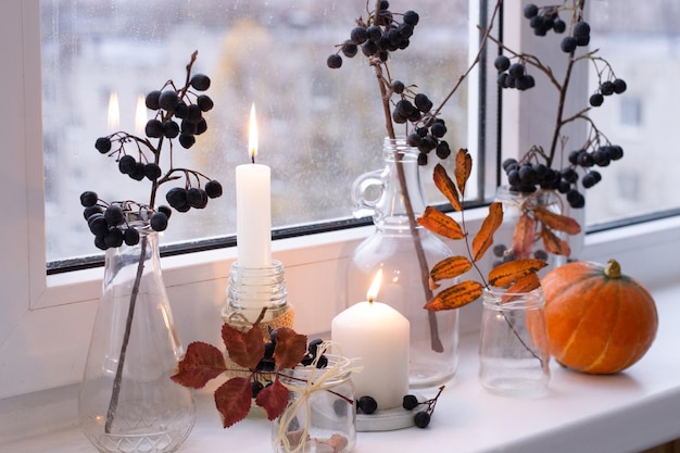 Autumn decoration of window. Branches of chokeberry, dry leaves in jars, pumpkin and candles.