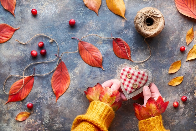 Foto decorazioni autunnali realizzate con materiali naturali, foglie autunnali luminose. le mani in maglione arancione tengono il cuore di legno. caduta stagionale laici piatta.