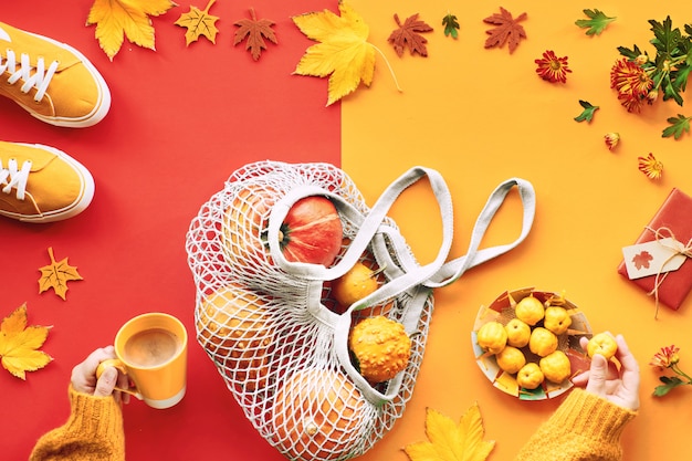 Autumn flat lay in green and orange with pumpkins in string bag, sneakers and female hands with cup of coffee and yellow fruit