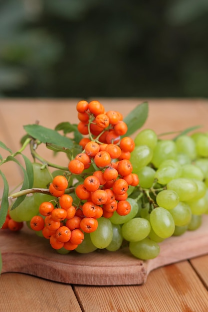 木製のテーブルのクローズアップの秋のブドウとナナカマドの果実ぼやけた背景の上の木の板に熟した緑のブドウとオレンジ色のナナカマドの果実